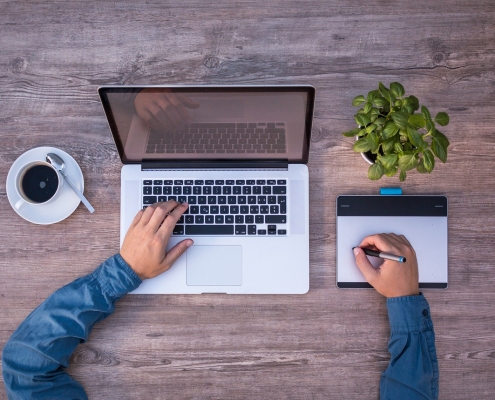 Foto: linke Hand liegt auf Laptop, rechte Hand hält Stift auf einem Grafiktablett